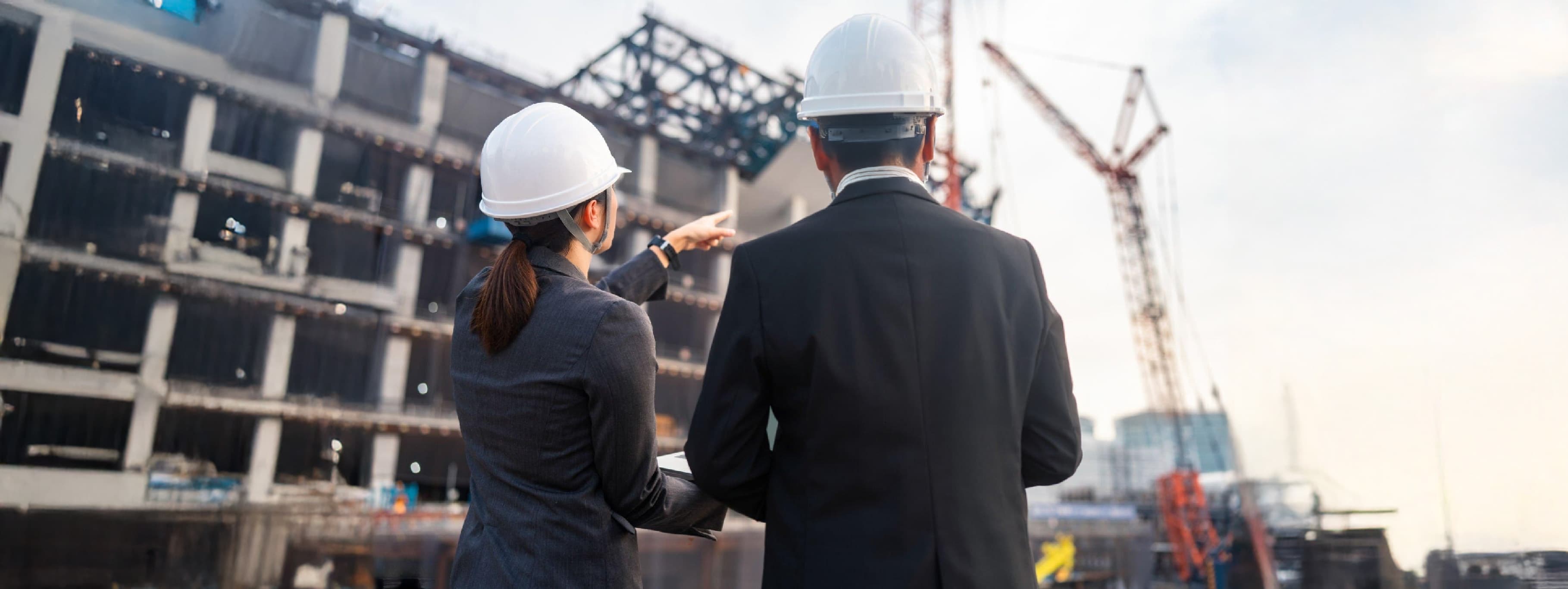 Deux ingénieurs en casque de chantier observent et discutent devant un immeuble en construction.